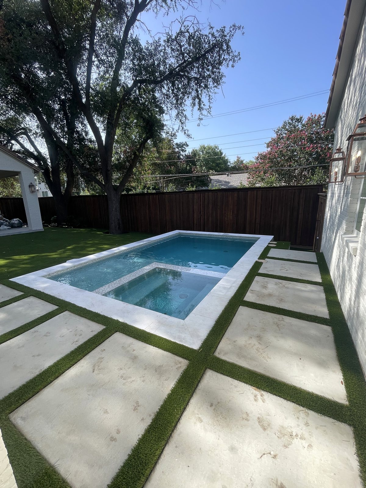 Full backyard view: rectangular pool with built-in spa, white stone coping, concrete pavers with turf joints and artificial lawn under mature trees โ completed by Infinity Group DFW in Dallas, Texas