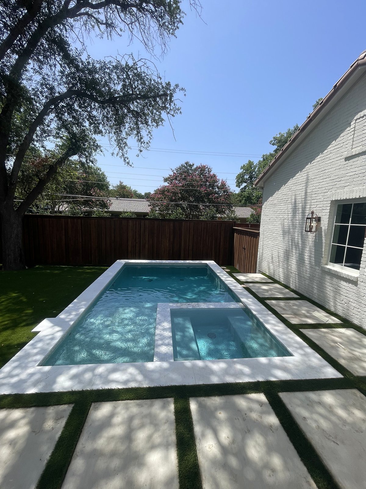 Pool and spa beside the white-brick home, framed by white coping and large pavers set in turf — completed by Infinity Group DFW in Dallas, Texas